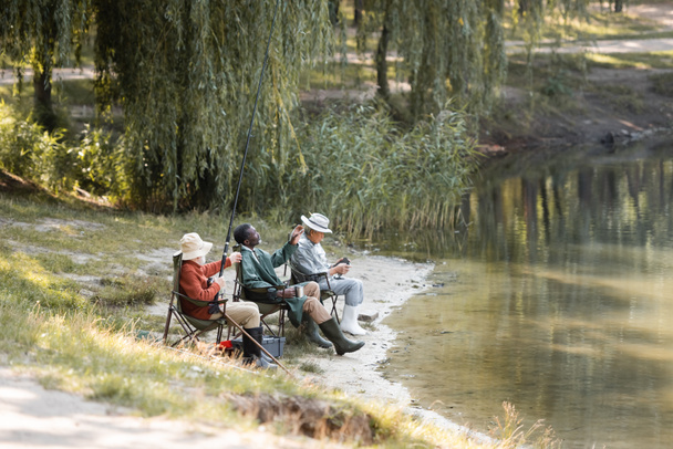 Multiethnic senior men with fishing rod sitting near lake in park  - Photo, Image