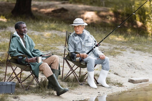 Asian man with fishing rod looking at african american friend with thermo cup near lake  - Photo, Image