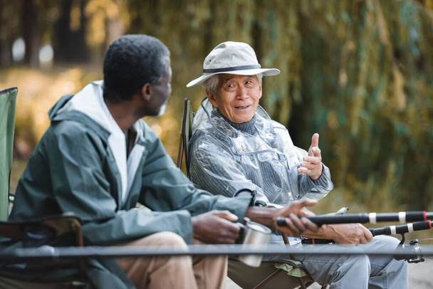 アフリカ系アメリカ人の友人と屋外で釣りをしながら話しているシニアアジア人男性  - 写真・画像