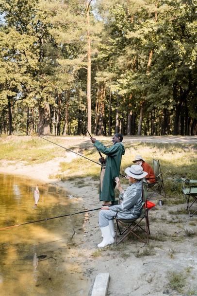 Мультикультурная рыбалка старших мужчин в озере рядом взволнованный друг в парке  - Фото, изображение