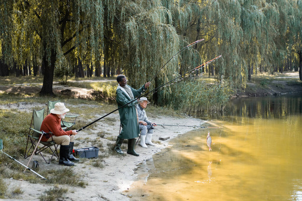 Hombre afroamericano captura de peces cerca de amigos interracial con cañas de pescar y lago  - Foto, imagen