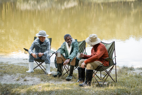 Interracial senior men in hats and fishing outfit holding thermo cups on chairs near lake  - Photo, Image