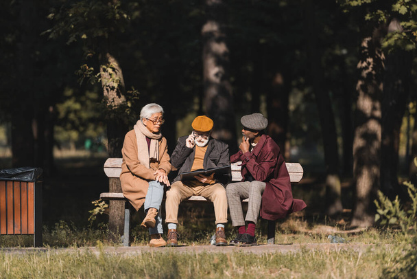 Старшие межрасовые мужчины сидят рядом с другом с фотоальбомом разговаривают на смартфоне в парке  - Фото, изображение