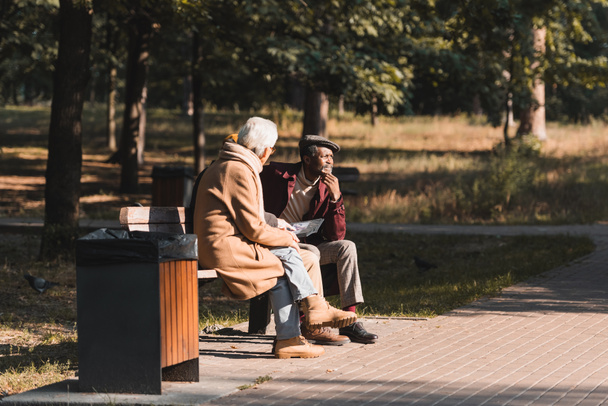 Uomo afroamericano seduto vicino agli amici con album fotografico nel parco durante l'autunno  - Foto, immagini