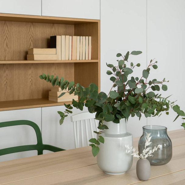 Close-up on simple and stylish vase with eucalyptus leaves on wooden dining table in room with white cabinets and wooden shelf - Foto, immagini
