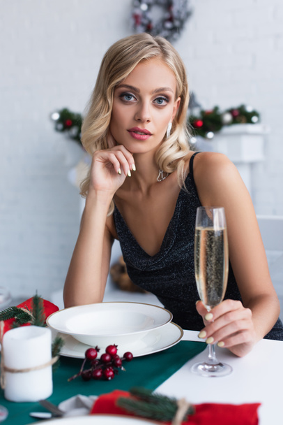 pretty and elegant woman looking at camera while sitting at festive table with glass of champagne - Foto, Imagen
