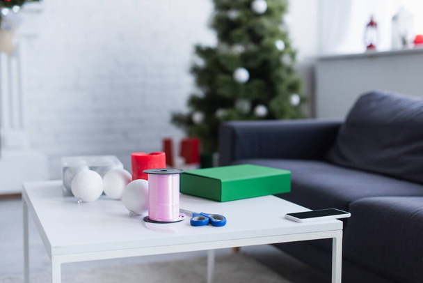 stůl s dárkovou krabičkou, cívkou s ozdobnou stuhou, cetkami a nůžkami v obývacím pokoji s rozmazaným vánoční stromečkem - Fotografie, Obrázek