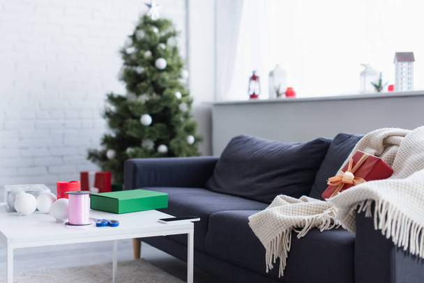 blanket and gift box on sofa near table with decorative ribbon and scissors in living room with blurred christmas tree - Photo, Image