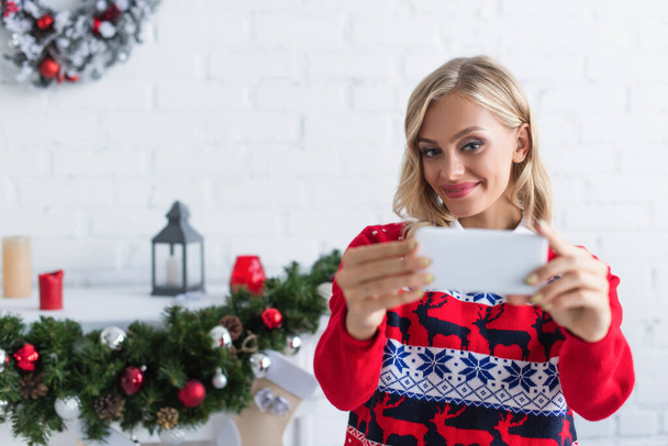 joyful woman in stylish sweater taking selfie on smartphone near blurred christmas garland - Photo, Image