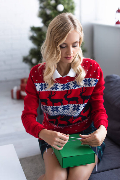 blonde woman in red sweater with ornament tying ribbon on green gift box - Photo, Image