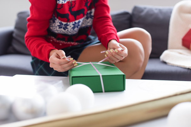vue recadrée d'une femme attachant un ruban décoratif sur une boîte cadeau près de boules de Noël floues - Photo, image