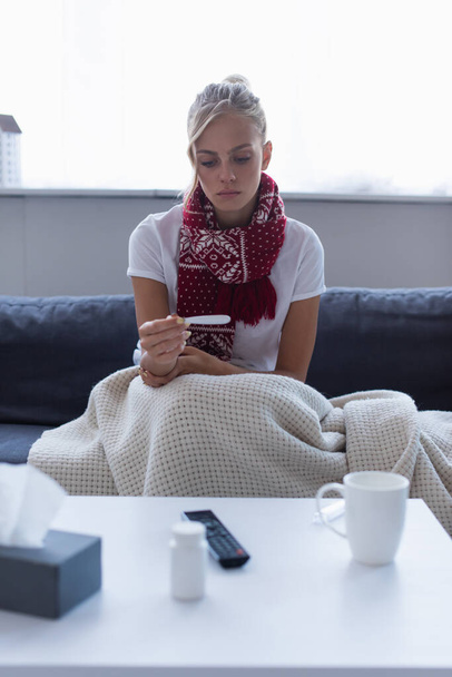 засмучена жінка в теплому шарфі дивиться на термометр поруч з розмитою тарілкою таблеток і теплим напоєм
  - Фото, зображення