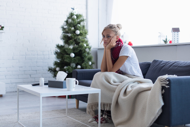 Kranke Frau sitzt auf Sofa unter warmer Decke neben verschwommenem Weihnachtsbaum - Foto, Bild