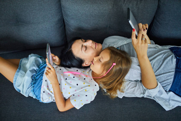 Mother and daughter browsing mobile phone while relaxing together on sofa at home, woman and girl play on smartphones while resting on couch on weekend day. - Foto, afbeelding