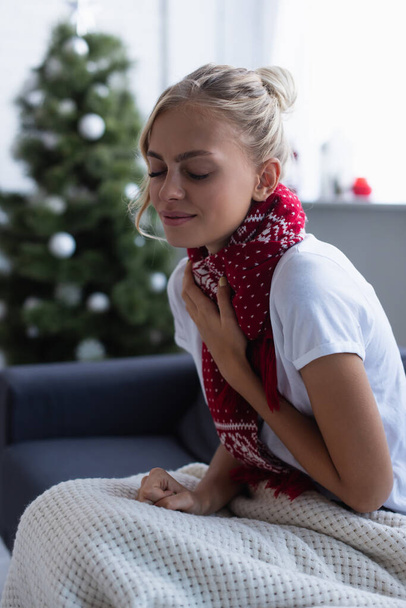 sick woman in warm scarf coughing on sofa with closed eyes near blurred christmas tree - Fotoğraf, Görsel