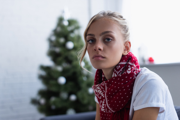 diseased woman in warm scarf looking at camera near blurred christmas tree - Photo, Image