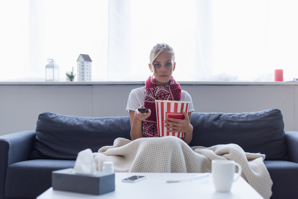 nemocná žena s dálkovým ovladačem a kbelík popcornu sledování televize v blízkosti rozmazané papírové ubrousky a šálek teplého nápoje - Fotografie, Obrázek