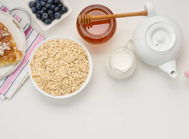 Ωμό πλιγούρι βρώμης σε λευκό κεραμικό πιάτο, βατόμουρο, μέλι σε λευκό τραπέζι, πρωινό. πάνω όψη - Φωτογραφία, εικόνα
