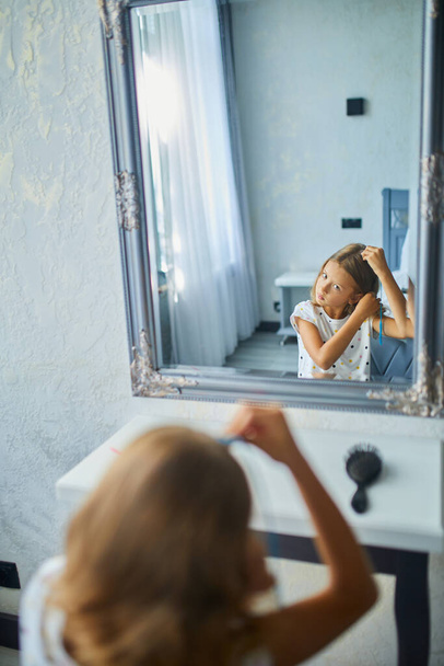 Ένα όμορφο κοριτσάκι να χτενίζεται μόνη της μπροστά από έναν καθρέφτη, να βουρτσίζει τα μαλλιά της στο σπίτι, να χαμογελάει, πρωινή ρουτίνα. - Φωτογραφία, εικόνα