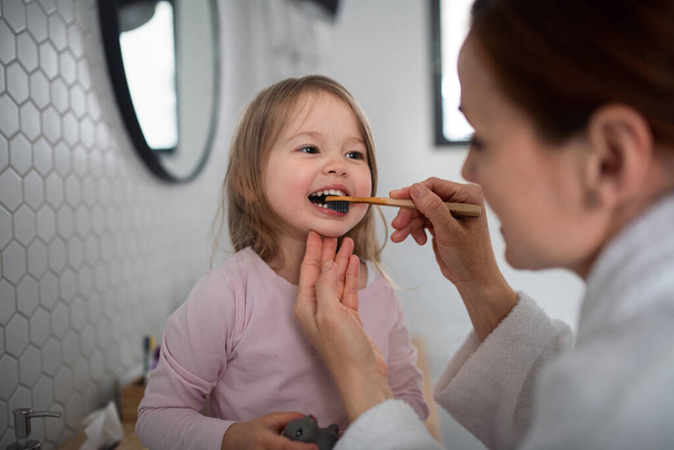 Mother with small daughter brushing teeth indoors in bathroom in the evening or morning. - Photo, image