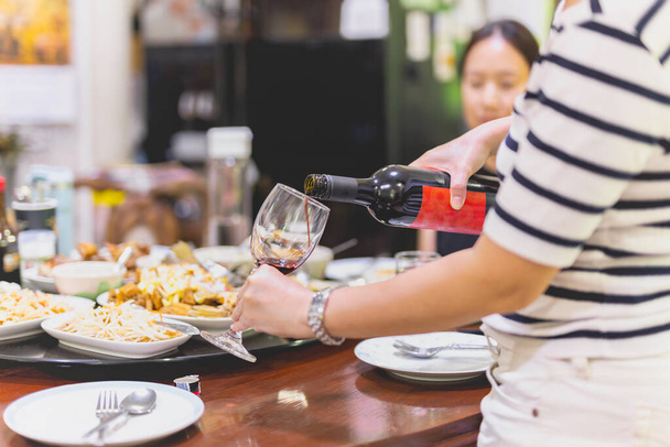 Χέρια γυναίκας ρίχνει κόκκινο κρασί σε δύο ποτήρια από το μπουκάλι με μια γυναίκα στο παρασκήνιο στο τραπέζι του δείπνου. - Φωτογραφία, εικόνα