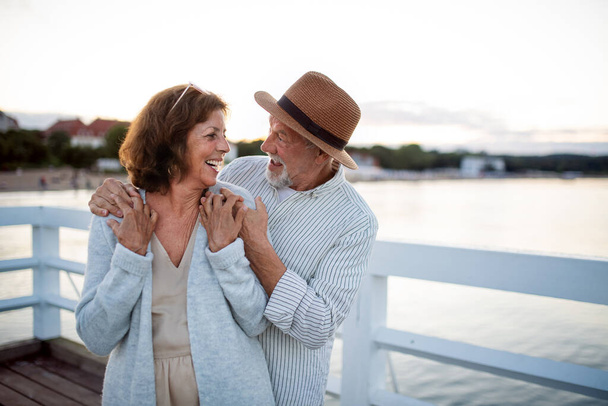 Happy senior couple hugging outdoors on pier by sea, looking at each other. - Фото, изображение