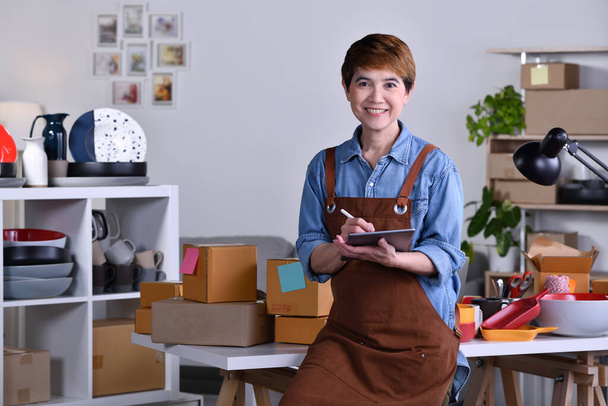 Madura mujer asiática emprendedora / propietaria de negocios de pie frente a su producto de cerámica de arcilla y trabajando en casa - Foto, Imagen