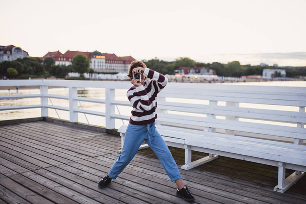 Portrait of preteen girl taking photo with camera on pier by sea at sunset, holiday concept. - Zdjęcie, obraz