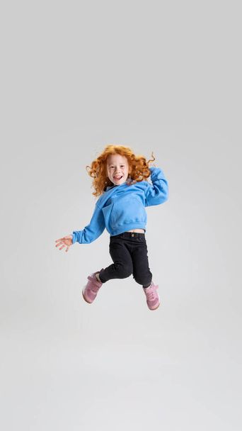 Portrait of little curly girl in blue sweater jumping isolated over gray studio background. Playing - 写真・画像