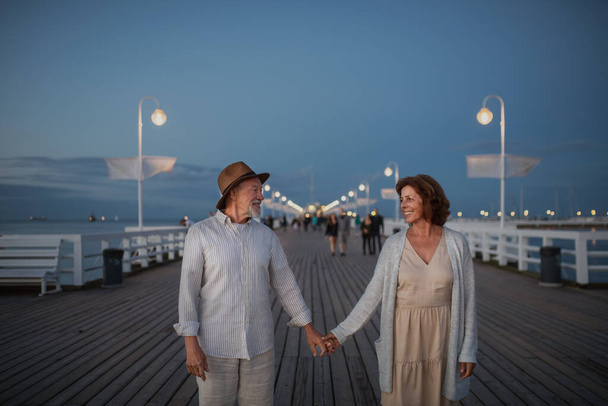 Front view of happy senior couple walking outdoors on pier by sea at dusk, holding hands. - 写真・画像