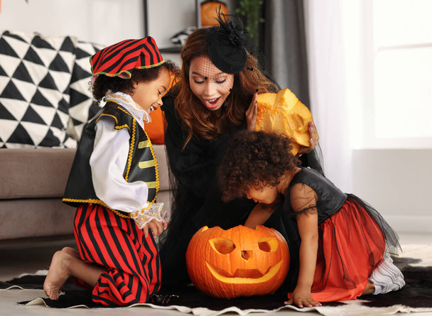 Gelukkig Afrikaans gezin moeder en kinderen dragen Halloween kostuums zitten op de vloer en kijken in een gesneden pompoen jack-o-lantaarn terwijl vieren All Saints Day samen thuis - Foto, afbeelding