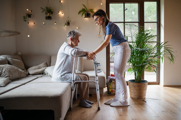 Side view of healthcare worker or caregiver visiting senior woman indoors at home, helping her to walk. - Foto, Bild