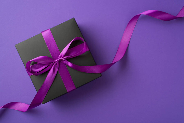 Top view photo of stylish black giftbox with violet satin ribbon bow on isolated violet background with blank space - Photo, Image
