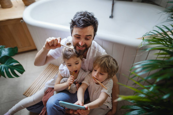 High angle view of father with two small children brushing teeth indoors at home and taking selfie, sustainable lifestyle concept. - Photo, image