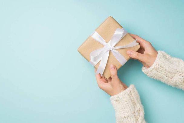First person top view photo of girl's hands in white sweater giving stylish craft paper giftbox with white satin ribbon bow on isolated pastel blue background with copyspace - Photo, Image