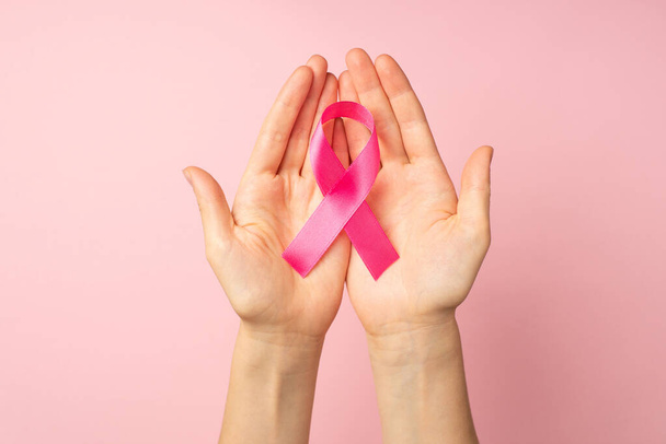 Πρώτη πρόσωπο top view φωτογραφία των χεριών κρατώντας ροζ κορδέλα στις παλάμες σύμβολο της ευαισθητοποίησης του καρκίνου του μαστού σε απομονωμένο παστέλ ροζ φόντο - Φωτογραφία, εικόνα