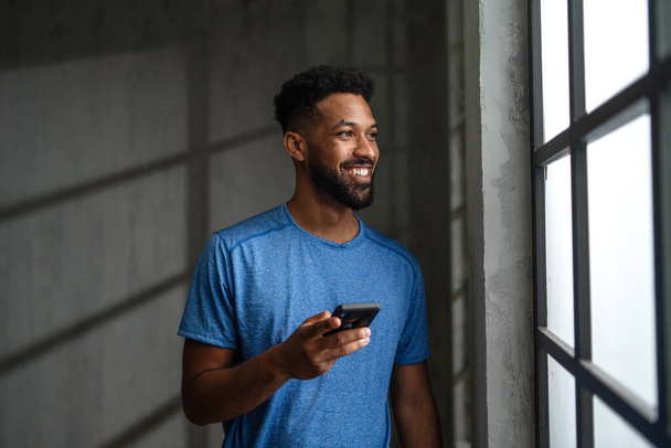 Happy young African American sportsman standing indoors at gym, using smartphone. - Photo, Image