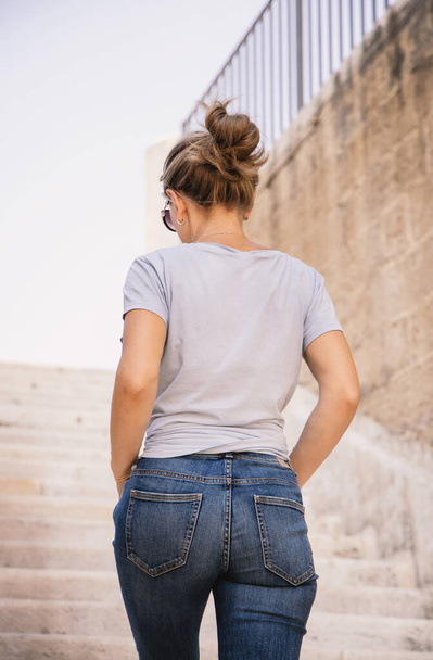 Young women wearing t-shirt and jeans going up the stone stairs in a sunny summer day, back view. T-shirt mockup . Three quarter sho - Foto, Bild