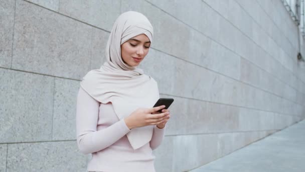Νεαρή μουσουλμάνα κυρία φορώντας μαντίλα δικτύωση σε smartphone, στέκεται σε εξωτερικούς χώρους, tracking shot, κενό χώρο - Πλάνα, βίντεο