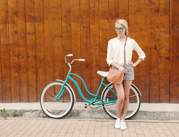 Young sexy blonde girl is standing near the vintage green bicycle with brown vintage bag in green sunglasses, warm, tonning - Fotó, kép