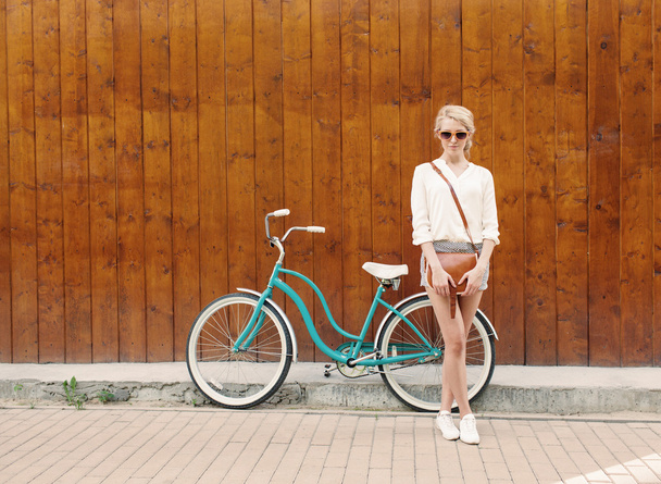 若いセクシーなブロンドの女の子は近くに立っているオレンジ色のサングラス、暖かい、茶色のビンテージ バッグ ビンテージの緑の自転車テニング - 写真・画像