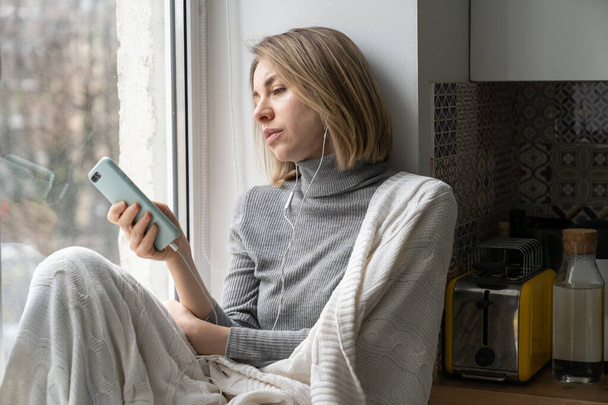 Aburrido mujer adicta y charlando en las redes sociales, sentado en el alféizar de la ventana en casa utilizando el teléfono móvil - Foto, Imagen