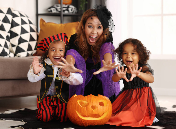 Joyful african american family mother and children in Halloween costumes making scary gesture and looking at camera while sitting on with jack-o-lantern in living room, celebrating all hallows day - Photo, Image
