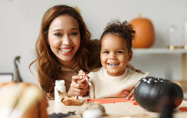 Krásná šťastná africká rodina matka a roztomilá holčička malování Halloween dýně společně doma, maminka a dítě sedí u stolu a připravuje ručně vyrobené dekorace pro všechny svatyně den - Fotografie, Obrázek