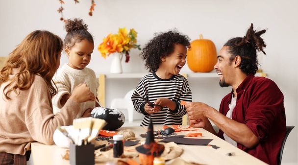 Joven familia afroamericana alegre madre, padre y dos niños preparándose para Halloween en casa, pintando calabazas y haciendo recortes de papel y decoraciones para la celebración del Día de los Santos - Foto, imagen
