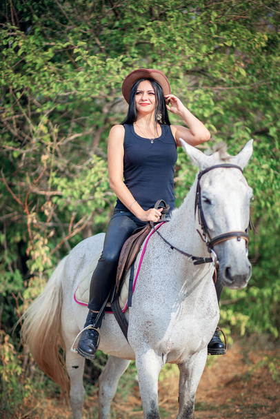 Una hermosa mujer en un sombrero de vaquero y pantalones de cuero monta un caballo gris. Entrenamiento a caballo - Foto, imagen