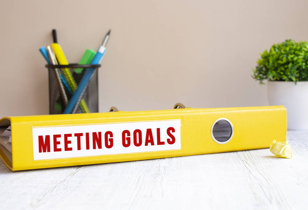 A yellow folder labeled MEETING GOALS is on the office desk. Flower and stationery background. Financial concept. - Photo, image