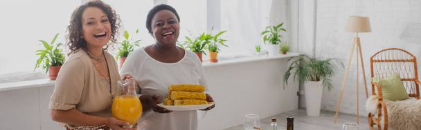 Positiva madre afroamericana e hija adulta sosteniendo jugo de naranja y maíz en casa, pancarta  - Foto, imagen