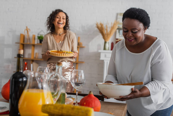 African american mother holding dish near thanksgiving dinner and blurred daughter at home  - Photo, Image