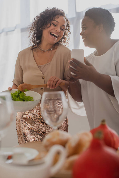 African american mother holding cellphone near cheerful daughter with salad and thanksgiving dinner  - Foto, Imagen
